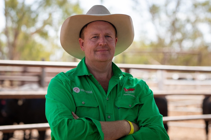 A man stands with arms crossed, wearing a bright green work shirt and cowboy hat. 
