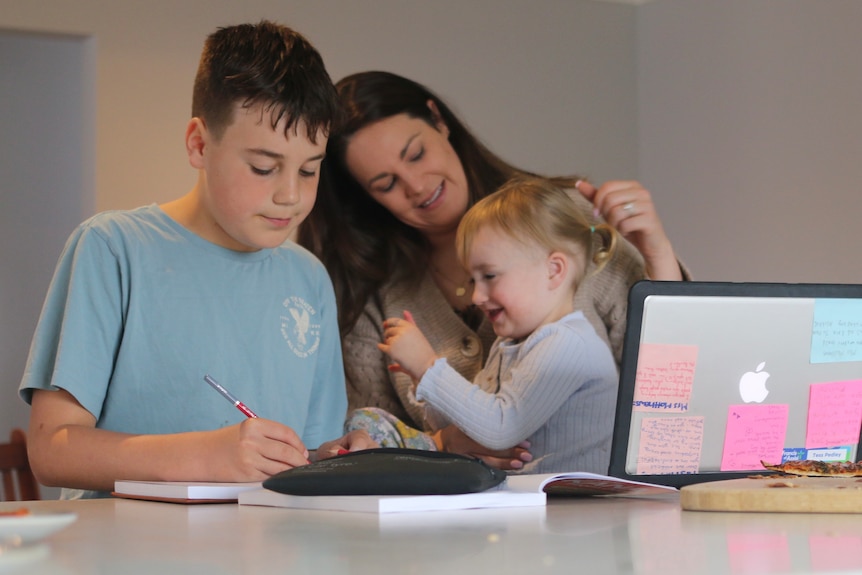 A young boy writing something at a table with his mum and baby sister nearby