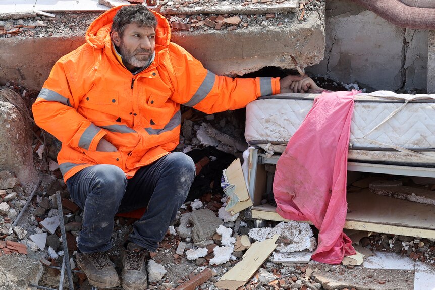 A man sits next to wreckage holding a hand that's under the rubble