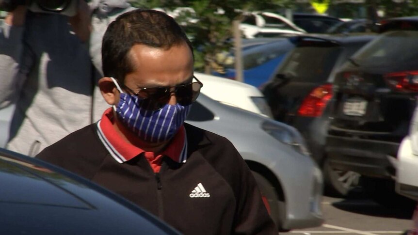 A man wearing big sunglasses and a blue and white face mask walks toward a car in a busy carpark