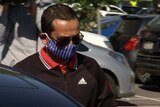 A man wearing big sunglasses and a blue and white face mask walks toward a car in a busy carpark