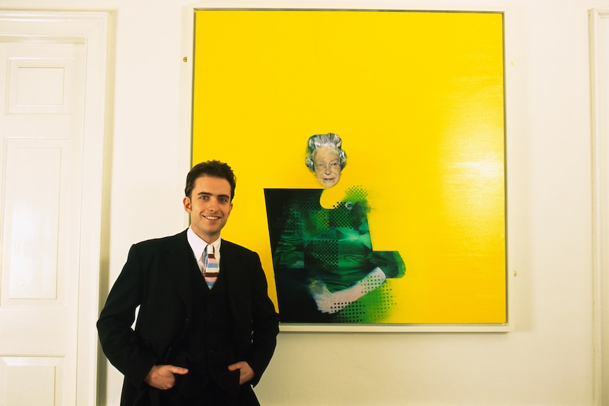 A young man in a suit stands smiling next to a surreal portrait of Queen Elizabeth II on a bright yellow background.