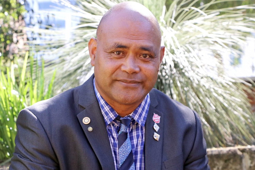 A head and shoulder shot of Moe Turaga in a suit, with shrubs in the background.