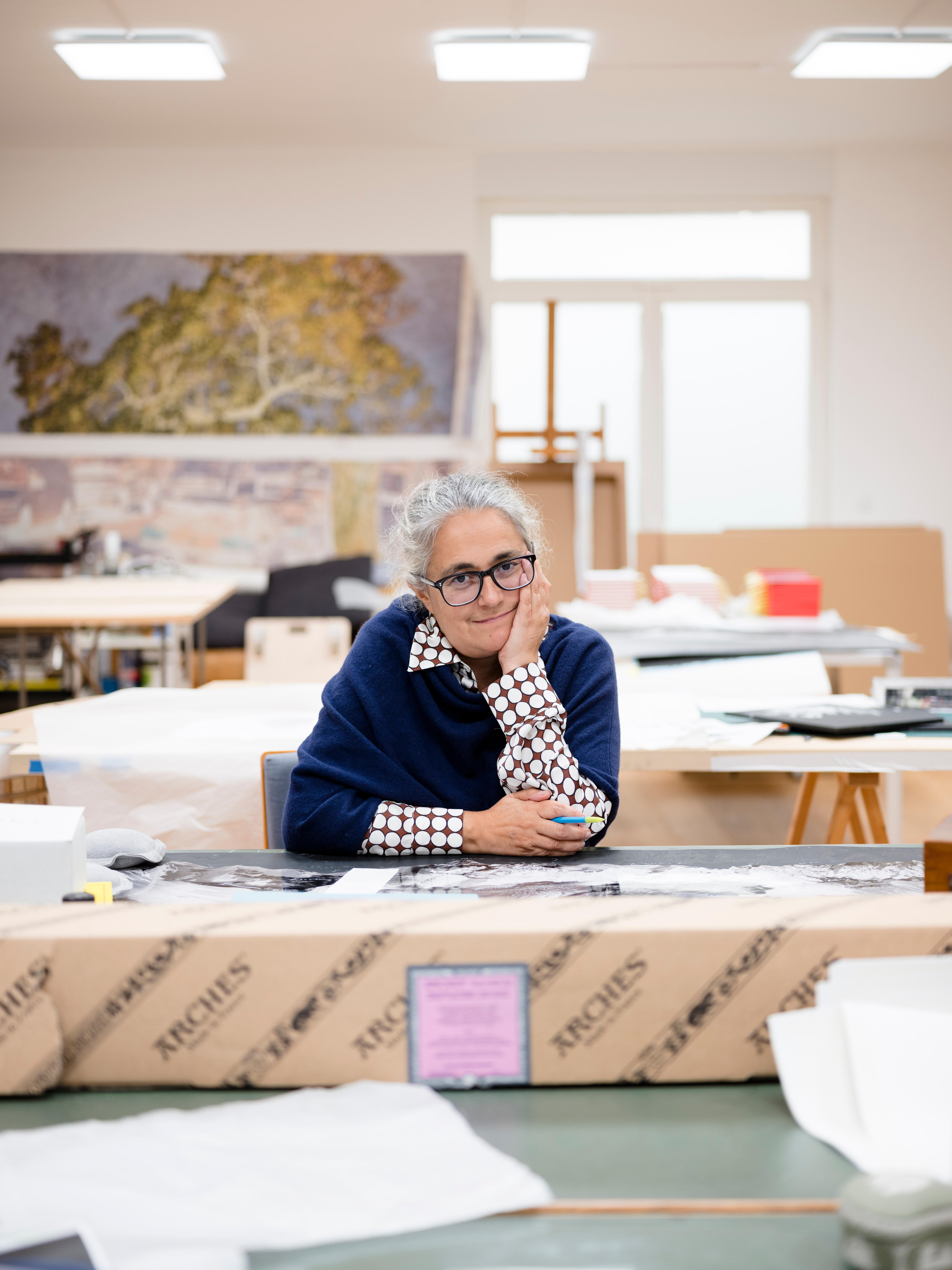 A grey-haired woman wearing black-rimmed glasses and a navy jumper sits in a cluttered room
