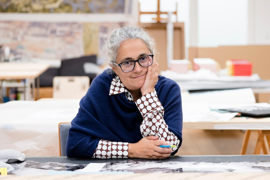 A grey-haired woman wearing black-rimmed glasses and a navy jumper sits in a cluttered room