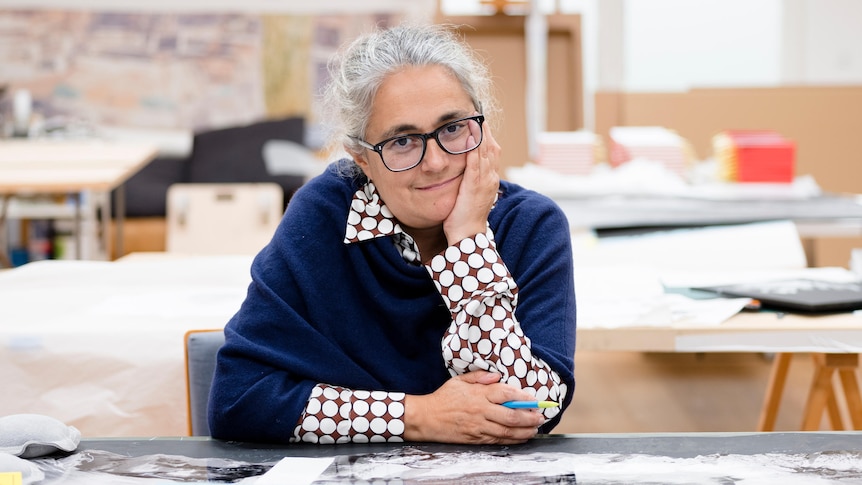 A grey-haired woman wearing black-rimmed glasses and a navy jumper sits in a cluttered room