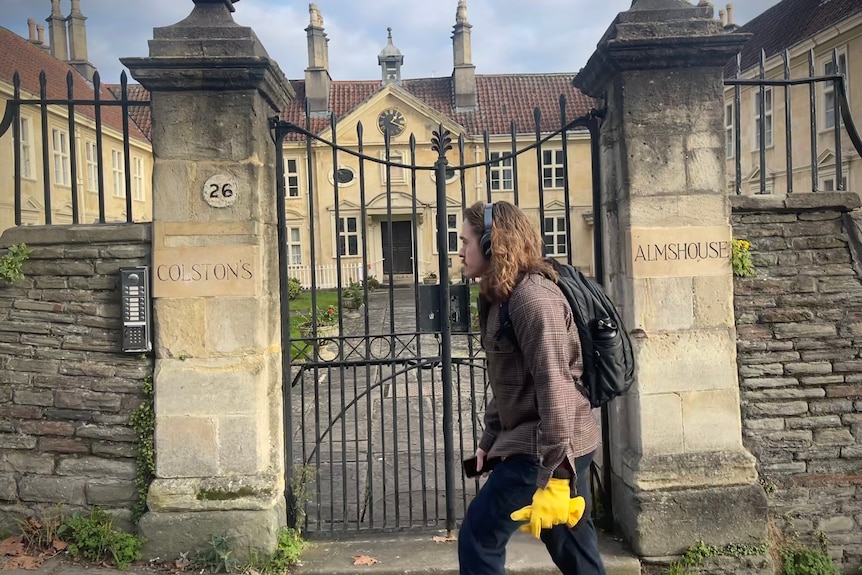 A man with long hair walks past an historic sandstone building with Colston Almshouse written on the gate