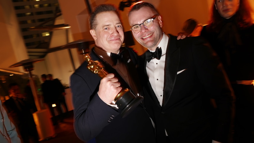 Brendan Fraser (left) and Samuel D. Hunter (right) pose for a photograph in a crowded room. Fraser is holding an Academy Award.