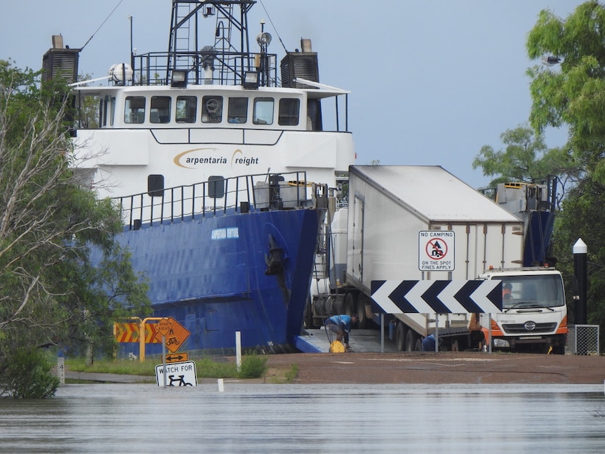 A large ship docked in a small inlet is unloaded by a truck amid floodwater