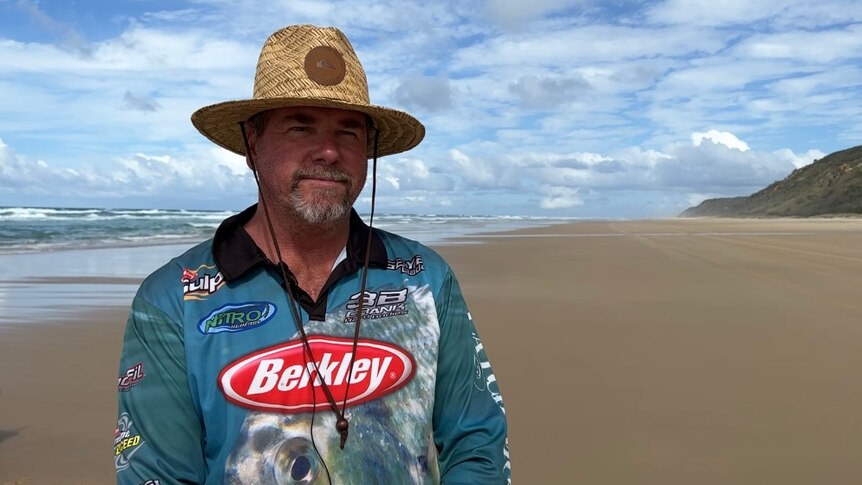 A man in a fishing shirt and hat stands on a beach