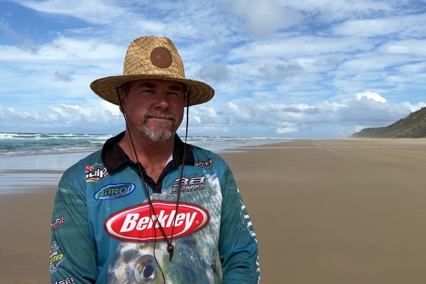 A man in a fishing shirt and hat stands on a beach