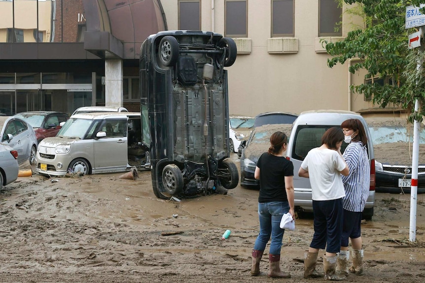 A car stands vertically among other damaged parked cars on a muddy road after being tipped over in a flood.