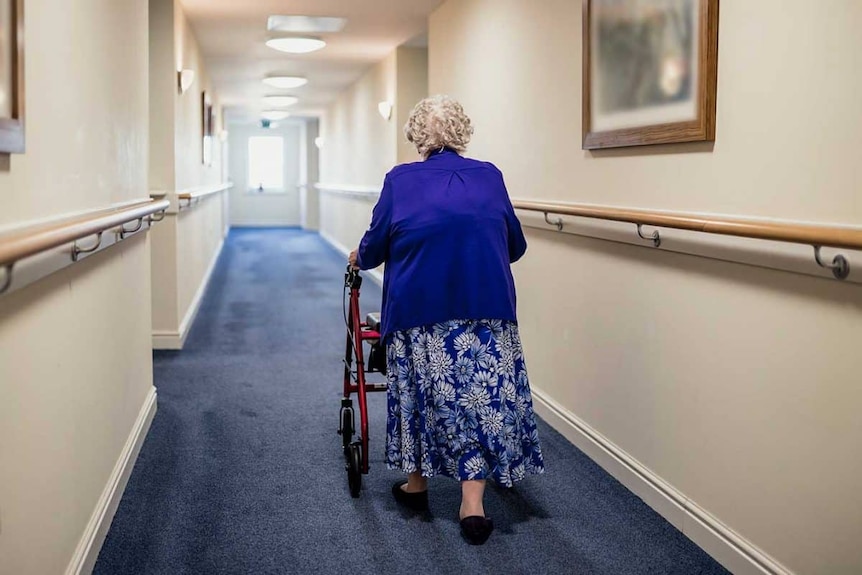 An elderly woman, seen from behind, makes her way down a hallway.