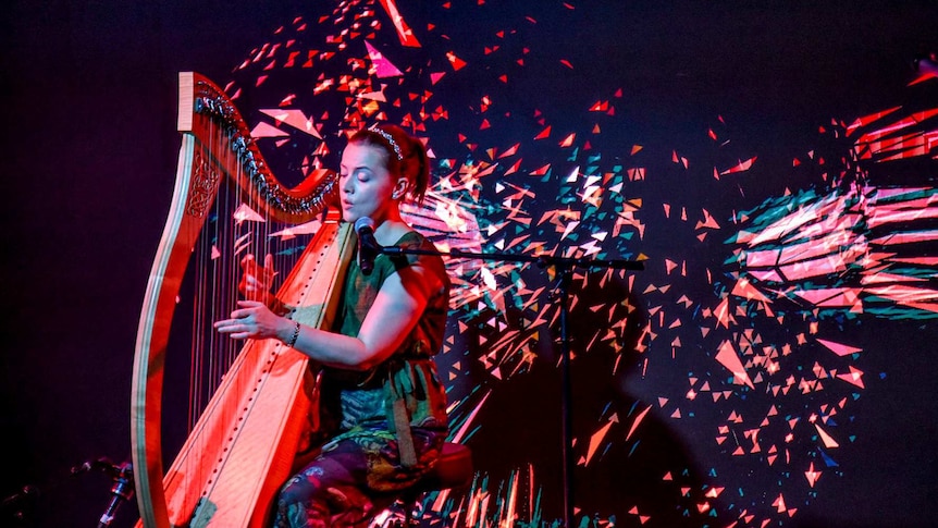 A woman plays a harp in front of a LED screen with fragmented colours.