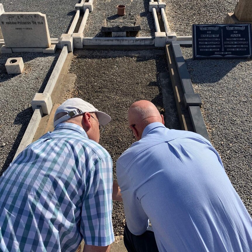 Two men leaning down to look at a grave in a cemetery.