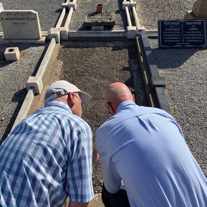 Two men leaning down to look at a grave in a cemetery.
