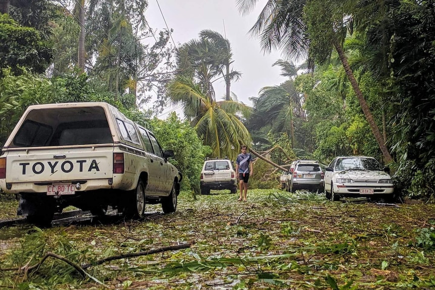 A man walks through a Darwin street littered with leaves, branches, and fallen trees.