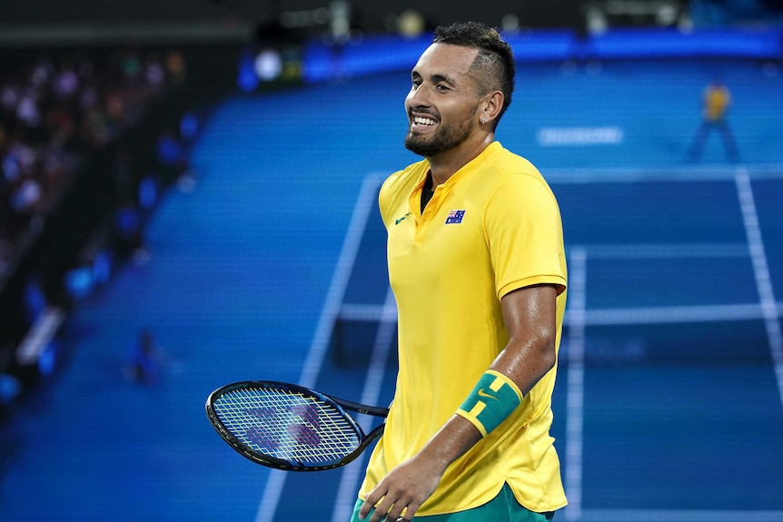 Nick Kyrgios smiles while on the court, holding his racquet. He's wearing green and gold.