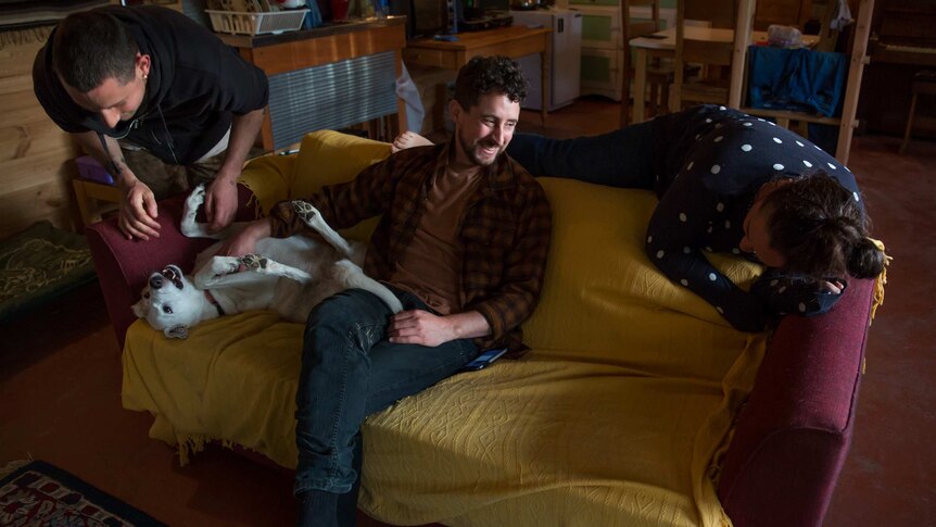 Three young adults laugh together as they relax on a couch with a dog.