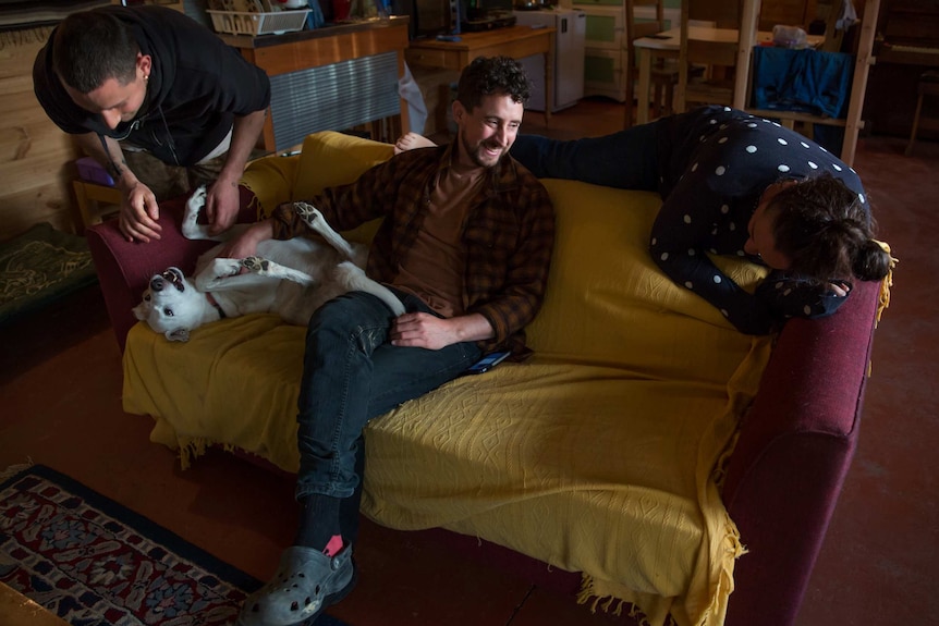 Three young adults laugh together as they relax on a couch with a dog.