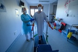 A man in a hospital gown, wearing a face mask, uses a treadmill with the help of a hospital physiotherapist.