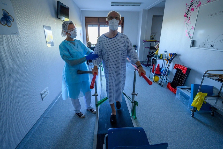 A man in a hospital gown, wearing a face mask, uses a treadmill with the help of a hospital physiotherapist.