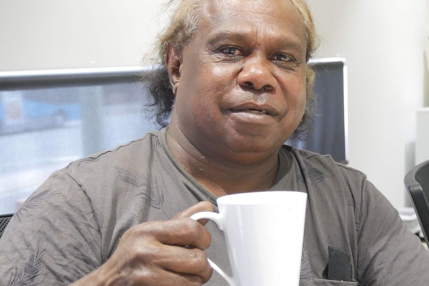 An Aboriginal man holds a mug of coffee in an indoor setting.