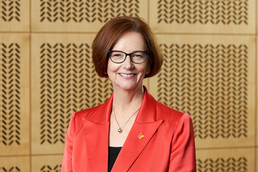 Julia Gillard in glasses and wearing a red jacket, standing in front of a patterned wooden wall