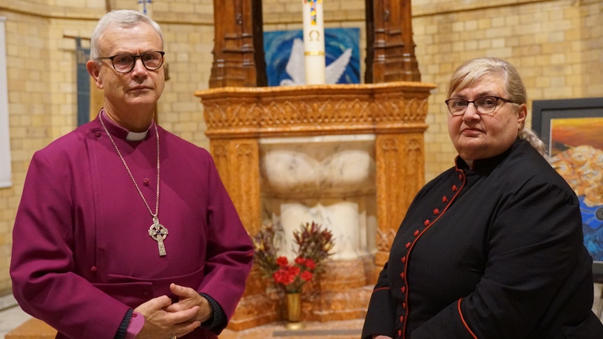 a priest wearing glasses and a woman reverend look at the camera