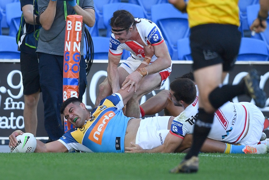 A Polynesian rugby player in light blue jersey stretches to score a try surrounded by opponents in white and red 