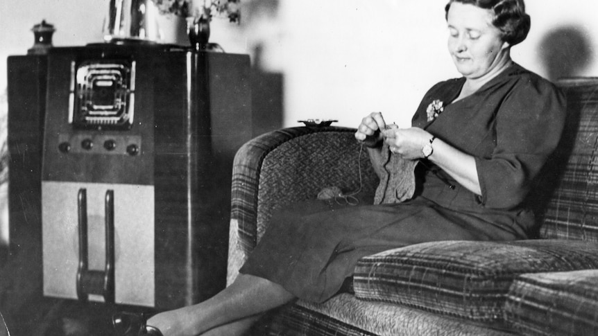 A woman in an armchair listens to the radio while knitting.