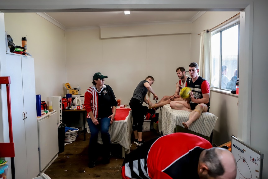 A football player has his ankle strapped in a white room with another player and support staff next to him