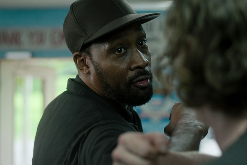 A man with a beard and cap faces another man, both have raised arms and clenched fists and stand facing each other in shop.