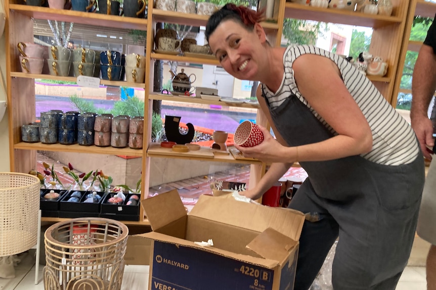 A woman in an apron bent over a carboard box in a shop, holding a cup