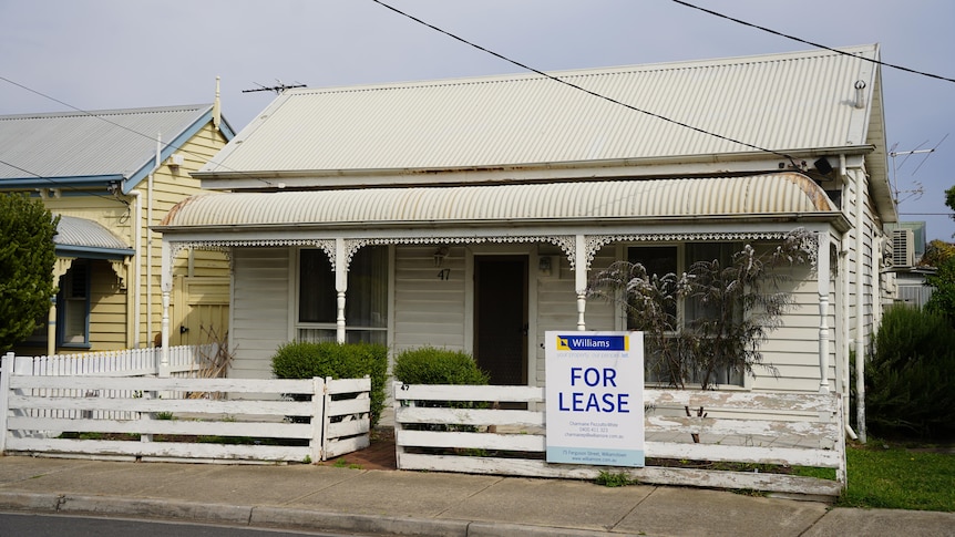 White weatherboard home with paint peeling off the fence and a for lease sign out the front