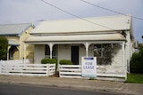 White weatherboard home with paint peeling off the fence and a for lease sign out the front