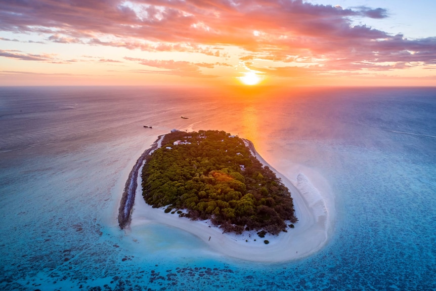 Drone shot of sun setting behind coral cay, vibrant colours, clear ocean.