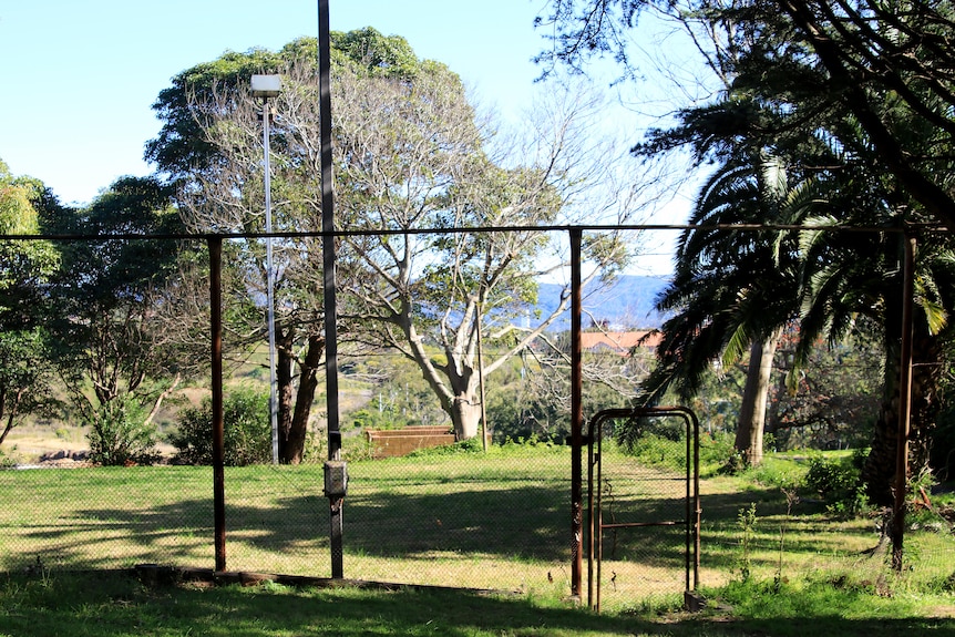 A rusted fence surrounds an unused lawn tennis court in a state of disrepair.