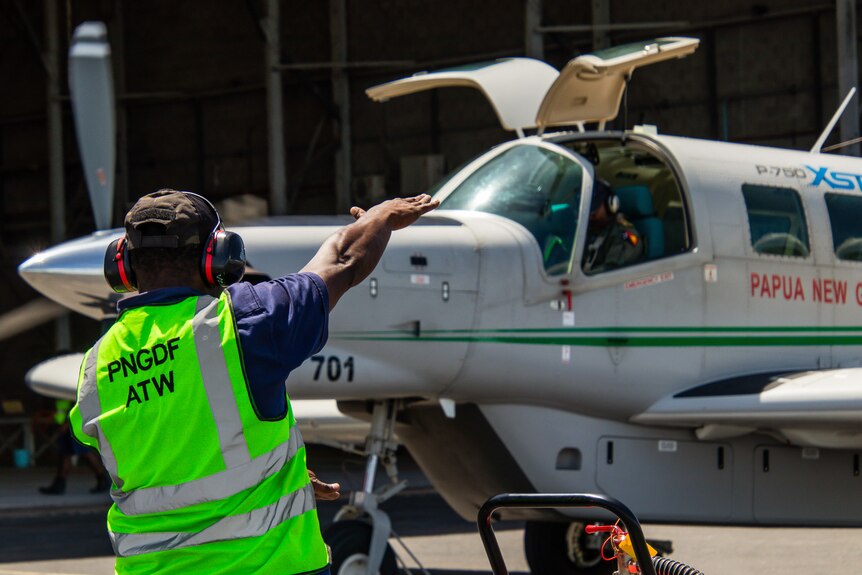 A man wearing a fluro vest and headphones flags down a plane with Papua New Guinea written on the side.