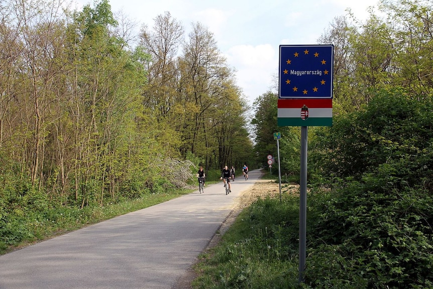 You view a narrow single-vehicle road in a verdant forested area with a sign carrying the EU and Hungarian flags.