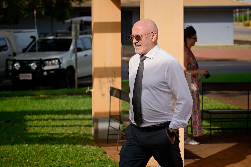 A man wearing a white shirt and black tie walks outside. He is serious.