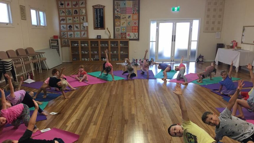 Children strike a yoga pose on mats placed in a circle in a hall.