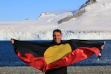 A man stands in front of a glacier holding the Aboriginal flag.