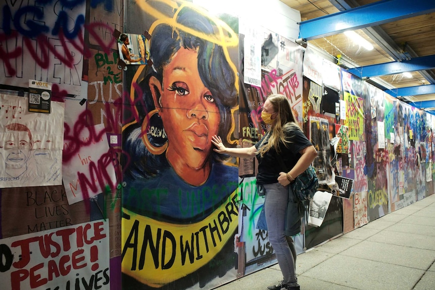 A woman in a face mask touches mural featuring Breonna Taylor's face