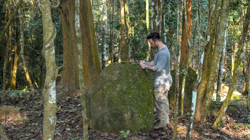 A man writes in a folder sitting on a boulder in thick jungle.