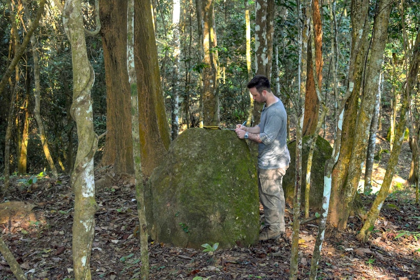 A man writes in a folder sitting on a boulder in thick jungle.