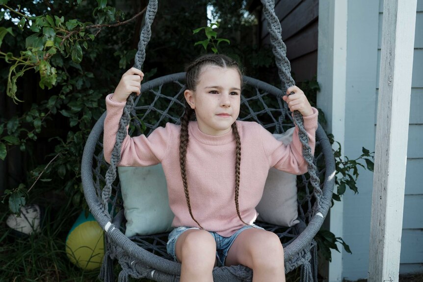 A little girl with long braids and wearing a pink sweater sits in a swing chair