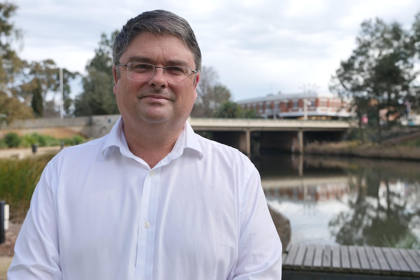 A man in a white shirt with glasses stands in front of a lagoon with a bridge going over it.