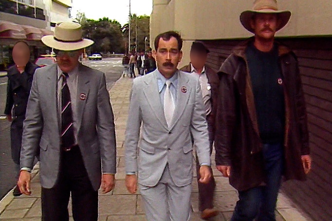 A short man with a moustache dressed in a grey suit walks along a footpath outside a court room