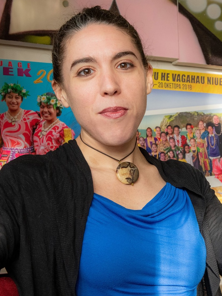 Woman in blue and black top looks at camera while leaning against wooden table.